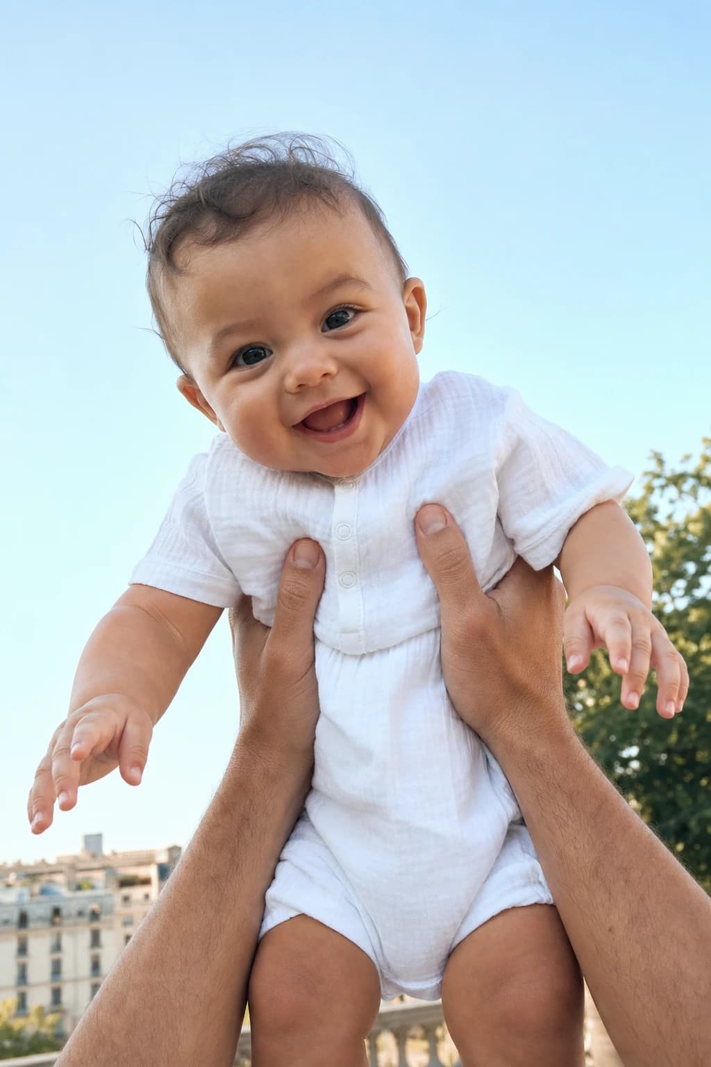 A smiling baby is held safely beneath a clear morning sky.